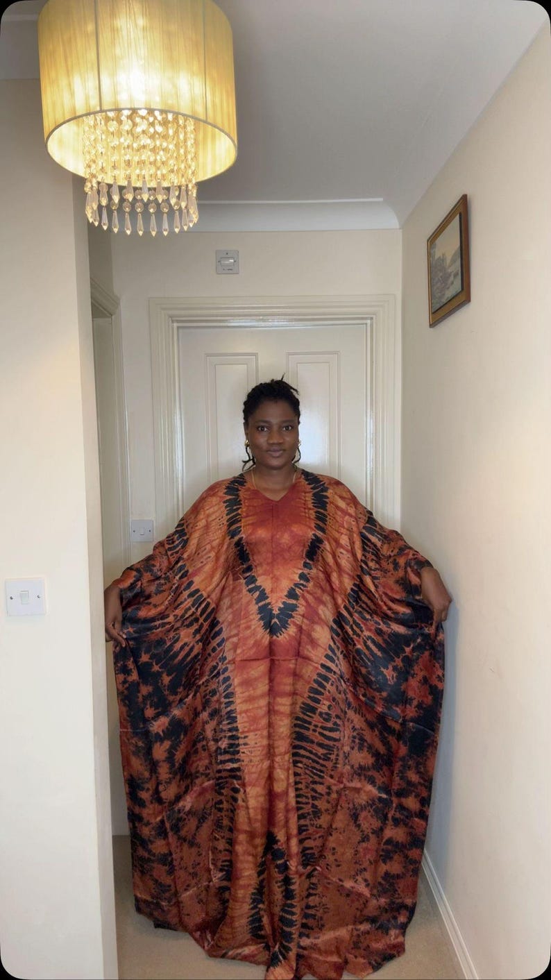 May include: A flowing, V-neck dress in shades of orange and brown with black tie-dye patterns. The dress has wide sleeves and a loose fit. The fabric appears to be lightweight and suitable for warm weather. The dress is worn by a person.