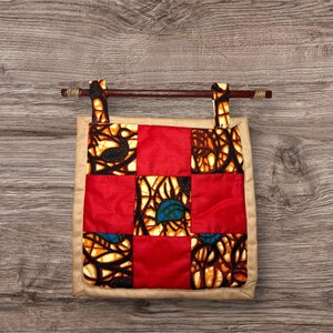 Puede incluir: Un adorno de pared cuadrado con un diseño de patchwork. El diseño presenta cuadrados rojos y cuadrados con un patrón negro, amarillo y naranja. El colgante está enmarcado con un borde beige y tiene una barra de madera en la parte superior para colgar.