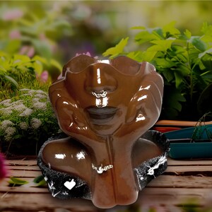 Op de afbeelding: Een bruine keramische gezichtspot met een glanzende afwerking. De pot toont een gedetailleerd gezicht met gesloten ogen, een neusring en een ketting. De handen ondersteunen het gezicht. De pot staat tegen een vage achtergrond van groen.