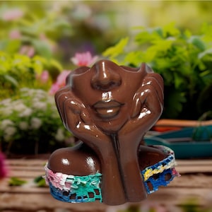 Op de afbeelding: Een bruine keramische gezichtspot met handen die de kin ondersteunen. De pot heeft een kleurrijke gehaakte kraag. Het gezicht heeft gesloten ogen en een serene uitdrukking. De achtergrond is een vage tuinscène.