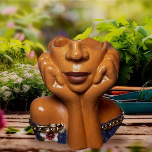May include: A brown ceramic planter shaped like a face, with hands supporting the chin. The planter has a smiling expression and is decorated with a gold chain and floral accents. The background is a blurred garden scene.