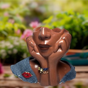 May include: A brown ceramic face planter with hands supporting the chin. The planter has a denim collar with a red lip pin and a gold and blue necklace. The background is a blurred garden scene.