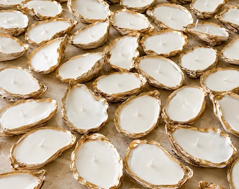 May include: A collection of numerous oyster shell candles, each filled with smooth white wax and featuring a single centered wick. The natural exterior of each shell is accented with a metallic gold painted rim, creating a uniform appearance.