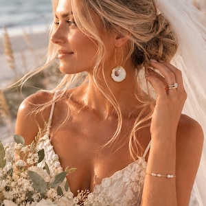 May include: A pair of gold hoop earrings featuring a circular white shell pendant with a small pearl accent, a delicate gold chain bracelet with three small pearls, and a gold ring with a central stone, worn with a white lace dress and veil.