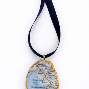 May include: A distinctive pendant with a map design set within a gold-edged oyster shell. It hangs from a navy blue ribbon. The map depicts the area around Perry, Florida. A stylish, handmade accessory.