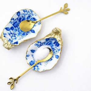 Könnte beinhalten: Zwei Austernschalen-förmige Schalen mit blauen Blumenmustern und goldfarbenen Rändern. Jede Schale enthält einen goldenen Löffel mit einem blattartigen Design am Griff. Eine Schale enthält weißes Salz. Auf einer weißen Oberfläche.