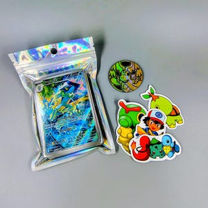 May include: A holographic plastic pouch containing a Frogadier trading card, accompanied by a circular holographic token and a collection of die-cut stickers featuring various animated characters including a trainer and several small creatures.