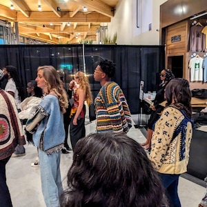 May include: A group of people are gathered, some wearing colorful, patterned jackets and sweaters. The setting appears to be an indoor event, possibly a fashion show or market, with clothing displayed on racks in the background.