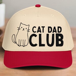 May include: A beige baseball cap with a red brim and button. The cap features a black outline drawing of a cat and the words "CAT DAD CLUB" in black, bold letters. The cap is a casual accessory.