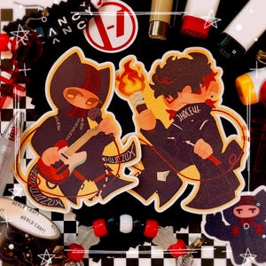 May include: Two stickers featuring cartoon figures. One plays a red guitar, the other holds a torch. Both wear dark outfits with text. The background includes pens, beads, and a black and white checkered pattern.