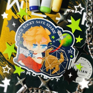 May include: A sticker featuring a cartoon illustration of a person with orange hair and glasses, with the text "GRACE ROCKY SAVE STARS!" and "PROJECT". The sticker is surrounded by star-shaped decorations and a silver chain.