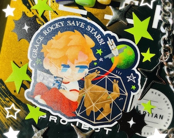 Op de afbeelding: Een sticker met een cartoon illustratie van een persoon met oranje haar en een bril, met de tekst "GRACE ROCKY SAVE STARS!" en "PROJECT". De sticker is omgeven door stervormige decoraties en een zilveren ketting.