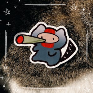 May include: A die-cut sticker depicting a cartoon character with a grey body, red scarf, and a red and green object extending from its face. The sticker is set against a background of dark fur with white decorative lines and stars.