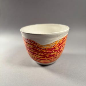 May include: A ceramic bowl with a white interior and rim. The exterior features a vibrant pattern of red and orange wavy lines against a white background. The bowl has a rounded shape and sits on a gray surface.