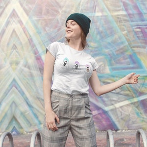 May include: A white t-shirt featuring three cartoon ghosts wearing hats in pastel colors. The person in the image is wearing a dark green beanie and plaid pants. The background is a colorful geometric mural.