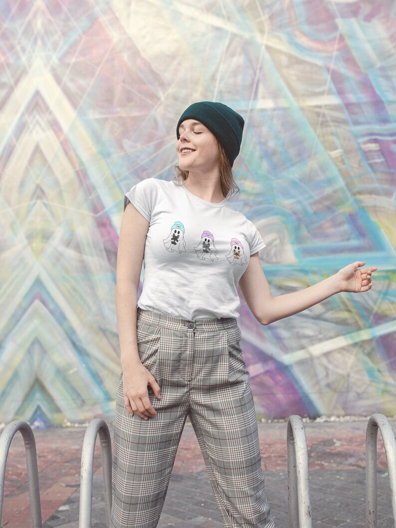 May include: A white t-shirt featuring three cartoon ghosts wearing hats in pastel colors. The person in the image is wearing a dark green beanie and plaid pants. The background is a colorful geometric mural.