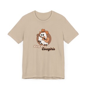 May include: Beige t-shirt featuring a cartoon ghost wearing a cowboy hat and playing a guitar. The ghost is inside a brown circle, with the words "Long Live Cowgirls" in brown script below the image.
