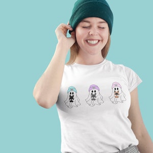 May include: White t-shirt featuring three ghost illustrations, each wearing a beanie and holding a small animal. The beanies are in blue, purple, and pink. The person in the image is wearing a green beanie.