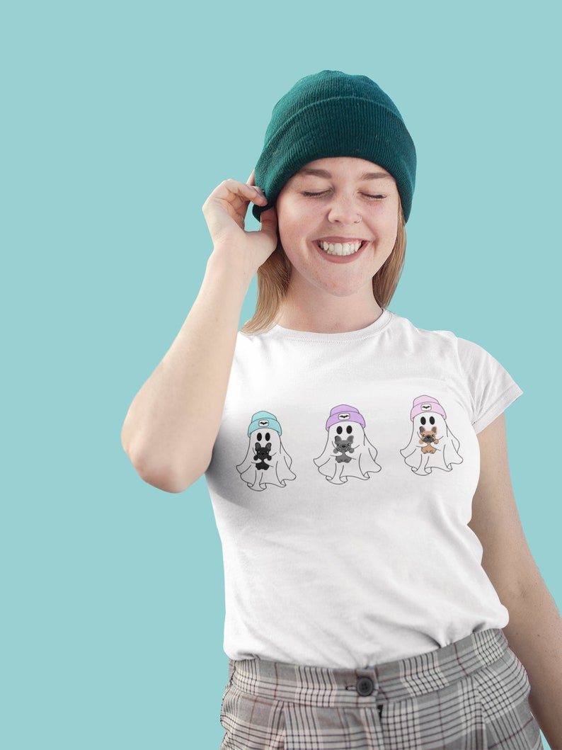May include: White t-shirt featuring three ghost illustrations, each wearing a beanie and holding a small animal. The beanies are in blue, purple, and pink. The person in the image is wearing a green beanie.