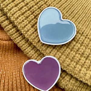 May include: Two heart-shaped pins with white borders. One is a gradient of blue, the other is purple. They rest on a folded, knitted, mustard-colored fabric. The background is a darker, brown-toned knit.