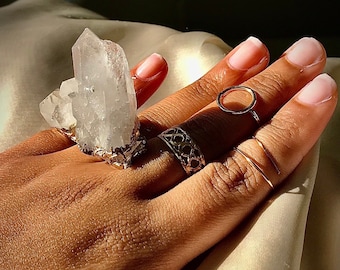 May include: A hand wearing a ring with a large clear quartz crystal cluster. The ring is on the index finger and there are two other gold rings on the middle finger.