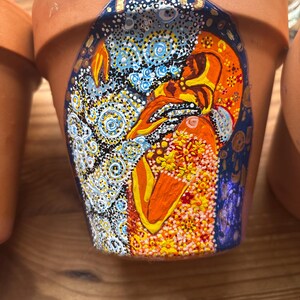 Könnte beinhalten: Terrakotta-Blumentopf mit handgemaltem Design. Das Design zeigt ein lebendiges, abstraktes Bild mit blauen, orangen, gelben und goldenen Farben. Der Topf ist unglasiert und hat eine einfache, klassische Form, geeignet für verschiedene Pflanzen.