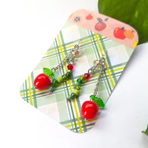 May include: Dangle earrings featuring a bright red apple charm with a green leaf, suspended from a silver-tone chain accented with small green and red beads. The earrings are displayed on a white card with a green plaid pattern and apple icons.