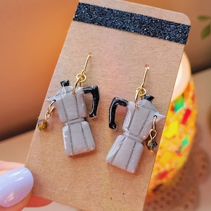 May include: A pair of handmade earrings featuring miniature gray coffee makers with black handles. The earrings have gold-colored hooks and small, dangling brown beads. The earrings are displayed on a brown card with a black glitter strip.