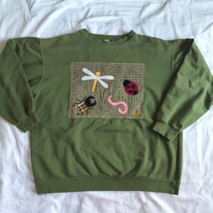 May include: An olive green crewneck sweatshirt featuring a rectangular patch on the chest. The patch displays fabric appliqués of a white and yellow dragonfly, a red and black ladybug, a brown plaid beetle, and a pink worm on a textured tan background.