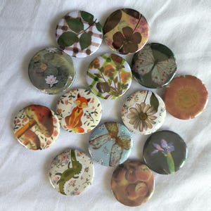 May include: A collection of round, decorative pinback buttons with various nature-themed designs. The buttons feature images of leaves, flowers, a fox, a butterfly, and other botanical elements. The color palette includes greens, browns, oranges, and blues.