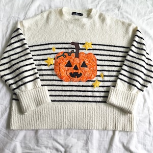 May include: A cream-colored textured knit sweater featuring horizontal black stripes. The center displays an orange pumpkin appliqué with a brown stem and black triangular facial features, surrounded by several small yellow fabric stars.