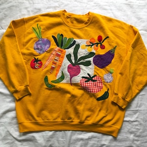 May include: Mustard yellow sweatshirt with an embroidered vegetable design. The design includes a purple onion, orange carrot, red tomato, green beans, and a purple eggplant. The vegetables are stitched onto a white square.