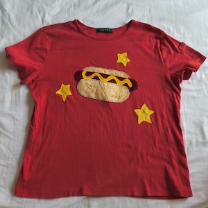 May include: A red short-sleeved t-shirt featuring a central fabric appliqué of a hot dog with yellow mustard, surrounded by three yellow fabric stars. The hot dog bun has the name "ang" embroidered in cursive script on the lower right side.