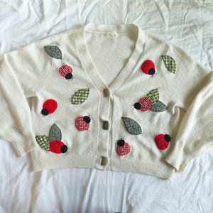 May include: Cream-colored cardigan sweater with a V-neck and button closure. The sweater is decorated with appliqued red ladybugs, black dots, and green leaves. The ladybugs have a checkered pattern. The sweater has long sleeves.