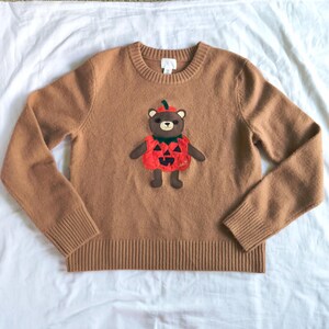 May include: A tan long-sleeve crewneck sweater featuring a central appliqué of a brown bear wearing a red pumpkin costume with a green stem on its head. The bear has black eyes, a nose, and a mouth, while the pumpkin has black triangular cutouts.