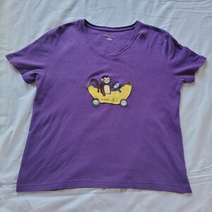 May include: Purple V-neck t-shirt with an embroidered design of a monkey riding a banana car. The banana car has wheels and the text "ang" is written on the banana. The shirt is made of a soft material.