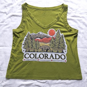 May include: Olive green tank top with a V-neck and a graphic of a bear, trees, mountains, and a sun. The word "COLORADO" is printed in white below the graphic.