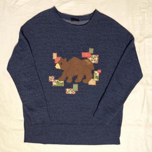 May include: A navy blue sweatshirt with a brown bear applique. The bear is surrounded by patchwork squares in red, green, and white patterns. The word "ang" is written on the bear's side.