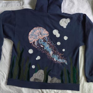 May include: Navy blue hooded sweatshirt featuring an embroidered jellyfish design. The jellyfish has a light blue and pink lace-like cap, with trailing tentacles. White bubbles and seaweed complete the underwater scene.