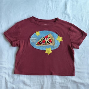 May include: A maroon short-sleeved t-shirt featuring a central embroidered patch. The patch displays a light blue oval background with a pizza slice, a small white cup, and three yellow stars. The pizza slice has red, black, and green toppings.