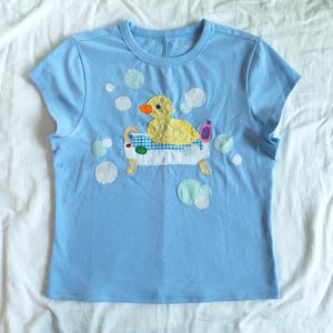 May include: Light blue t-shirt featuring an appliqued design of a yellow duck in a bathtub. The duck has an orange beak and is surrounded by white and light blue bubble accents. A pink bottle is also in the tub.