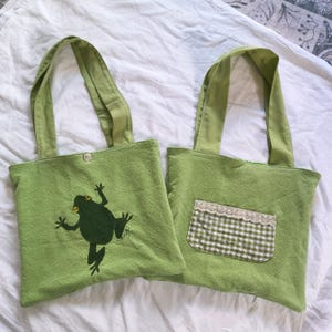 May include: Two light green tote bags. One bag features an embroidered dark green frog design. The other bag has a small pocket with a green and white checkered pattern and lace trim. Both bags have matching green handles.