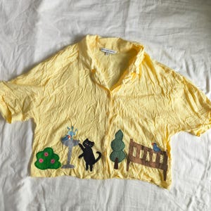May include: Yellow short-sleeved button-up shirt with a whimsical garden scene applique. The applique features a black cat, a bird, a tree, a bush, and a bench. The shirt has a collared neckline and a relaxed fit.
