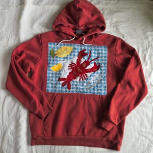 May include: Red hooded sweatshirt with a front pocket and a lobster applique. The applique features a red lobster, lemon slices, and a blue and white checkered background. The hoodie has a drawstring.