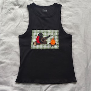 May include: A black sleeveless tank top featuring a rectangular fabric patch sewn onto the center. The patch displays a green and white gingham background with an embroidered ladybug playing a banjo next to a small orange campfire.