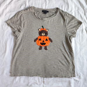 May include: A short-sleeved t-shirt with a cream and black horizontal striped pattern. The center features an applique of a brown bear wearing an orange pumpkin costume with a green stem and black triangular eyes and mouth. The label reads Sanctuary.