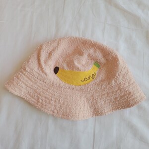May include: A light pink bucket hat with a yellow banana applique. The banana has a brown spot and a green tip, with the word "ang" embroidered in yellow. The hat is made of a textured fabric.