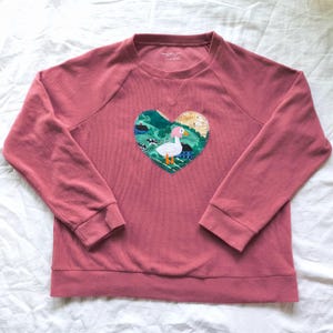 May include: Dusty rose-colored sweatshirt with a heart-shaped appliqué. The appliqué features a white goose with a pink head and orange feet, set against a landscape of green hills, blue water, and small houses. The sweatshirt has long sleeves and a crew neck.