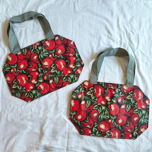 May include: Two tote bags featuring a vibrant red apple and green leaf pattern on a dark background. Each bag has a hexagonal shape and two grey and white gingham check fabric handles attached at the top with a small, centered metal button.