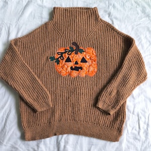 May include: A tan ribbed knit sweater featuring a high mock neck and long sleeves. Centered on the chest is an orange pumpkin appliqué with a patterned fabric texture, dark green leaves, and black triangular eyes, nose, and mouth details.